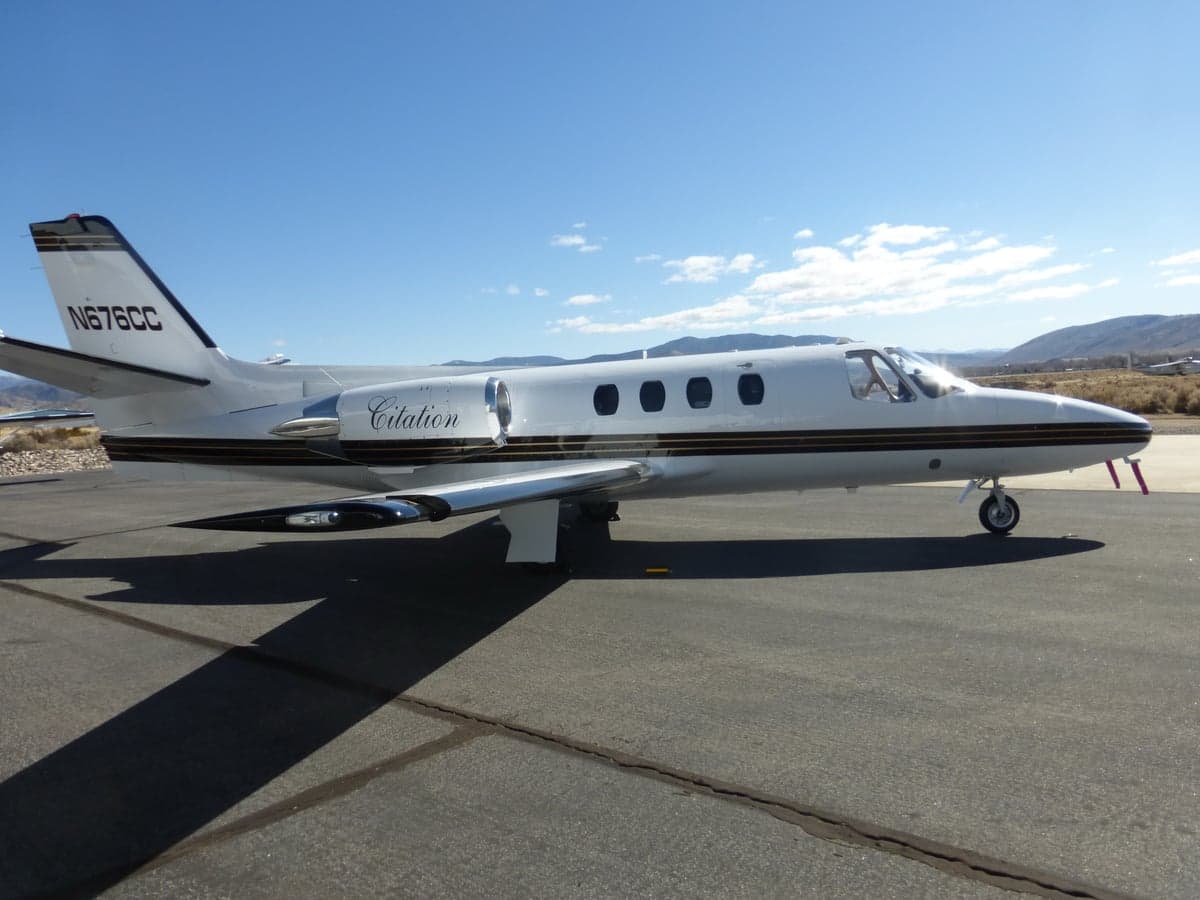 A white Cessna Citation jet with black and gold stripes, registration N676CC, parked on an airfield with mountains in the background.