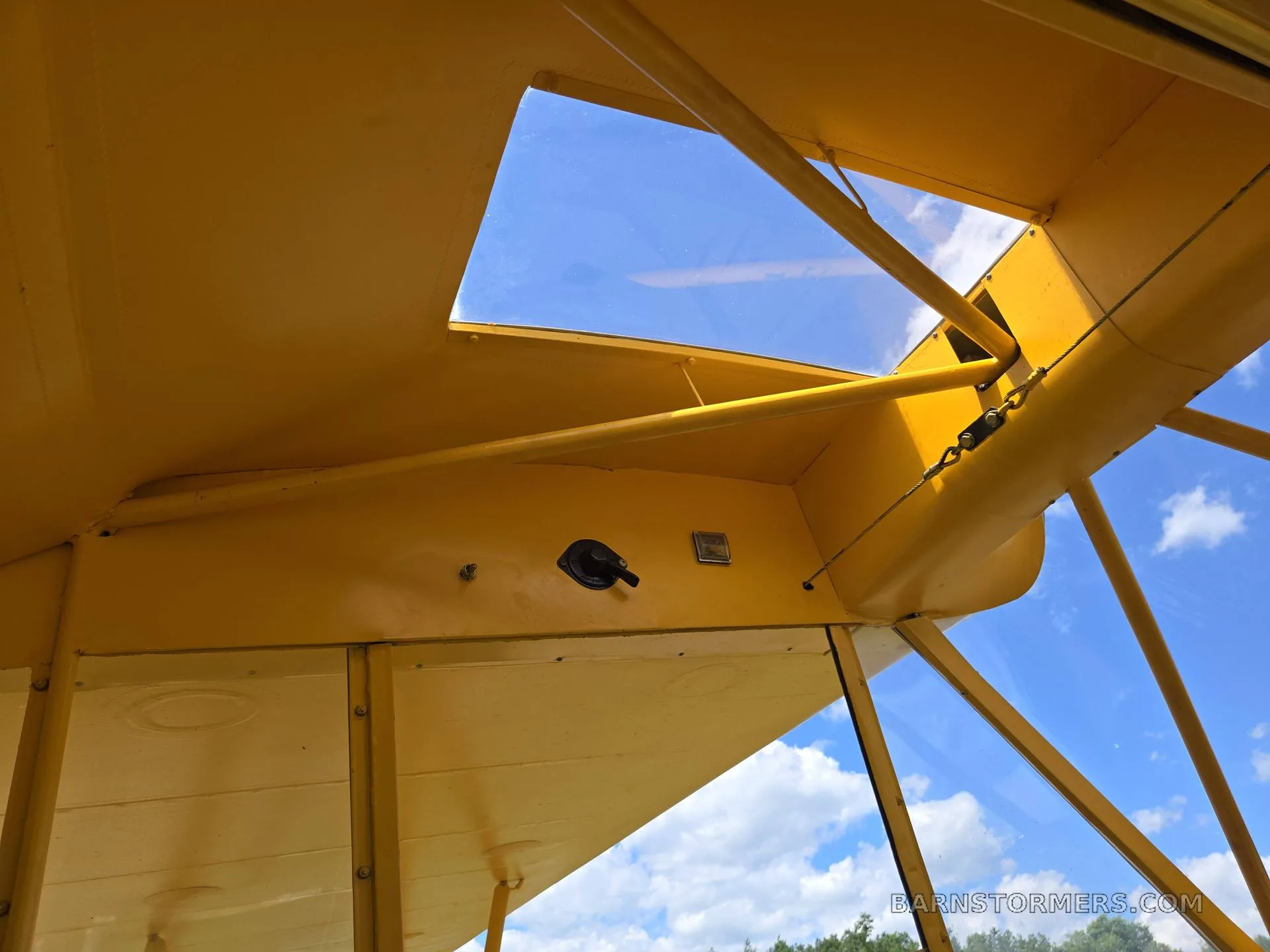 An upward view from inside a yellow aircraft, showing the wing structure, ceiling, and blue sky through a skylight.