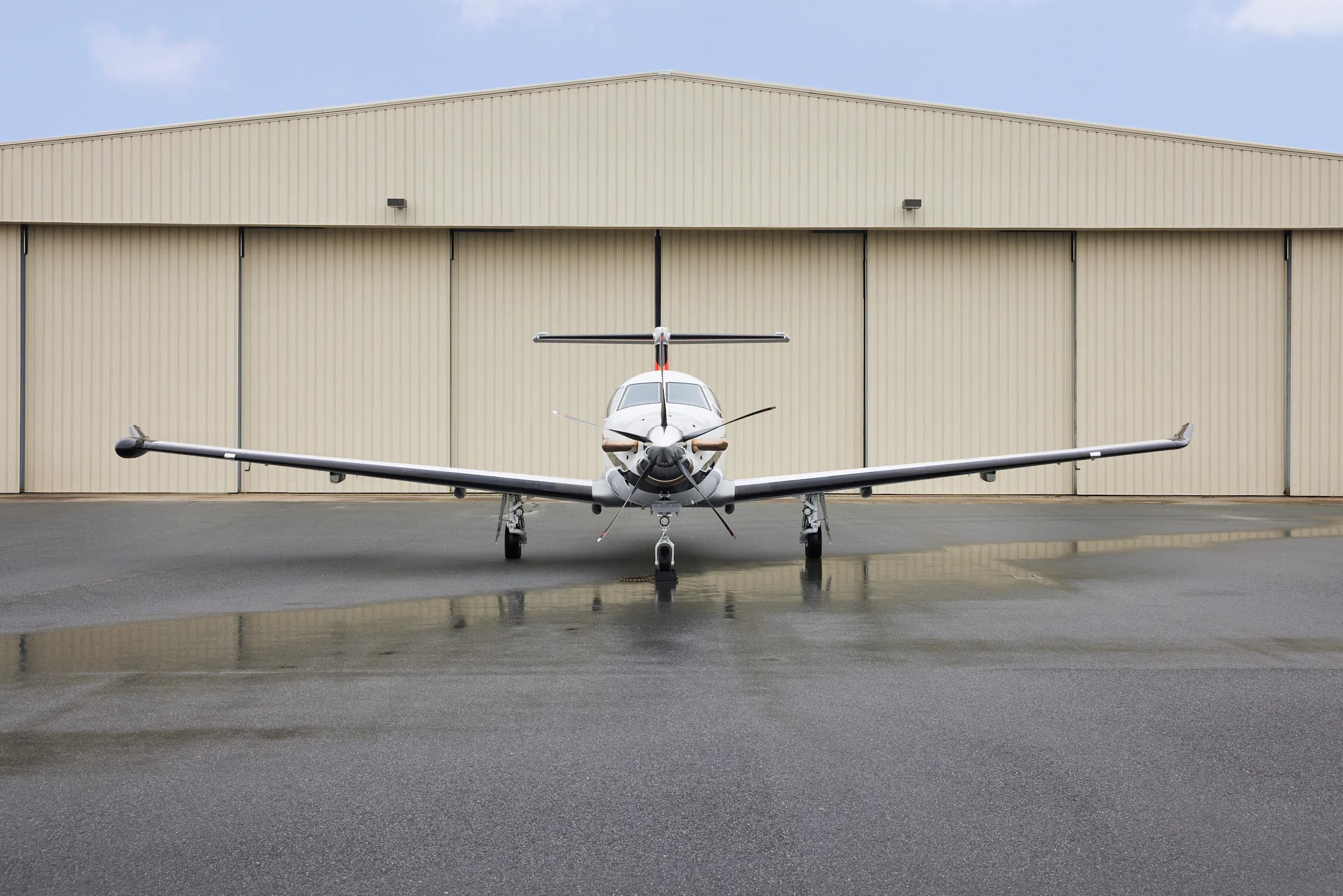 A white Pilatus PC-12 aircraft with black and orange accents parked head-on on a wet tarmac in front of a hangar.