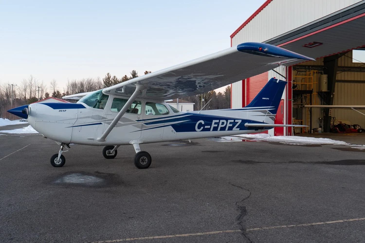 A white and blue Cessna 172 (C-FPFZ) is parked on an asphalt apron next to a hangar with some snow on the ground.
