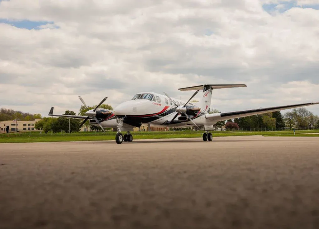 A white Beechcraft King Air 350 with black and red accents is parked on an airfield under a cloudy sky, viewed from a low angle.