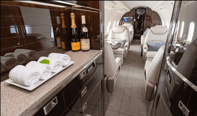 The galley area of a private jet features a counter with champagne bottles and rolled towels, leading into the passenger cabin.