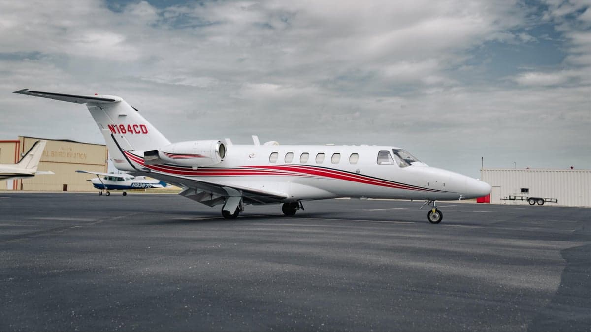 A white Citation CJ2 aircraft with red stripes is parked on the tarmac in front of a hangar.