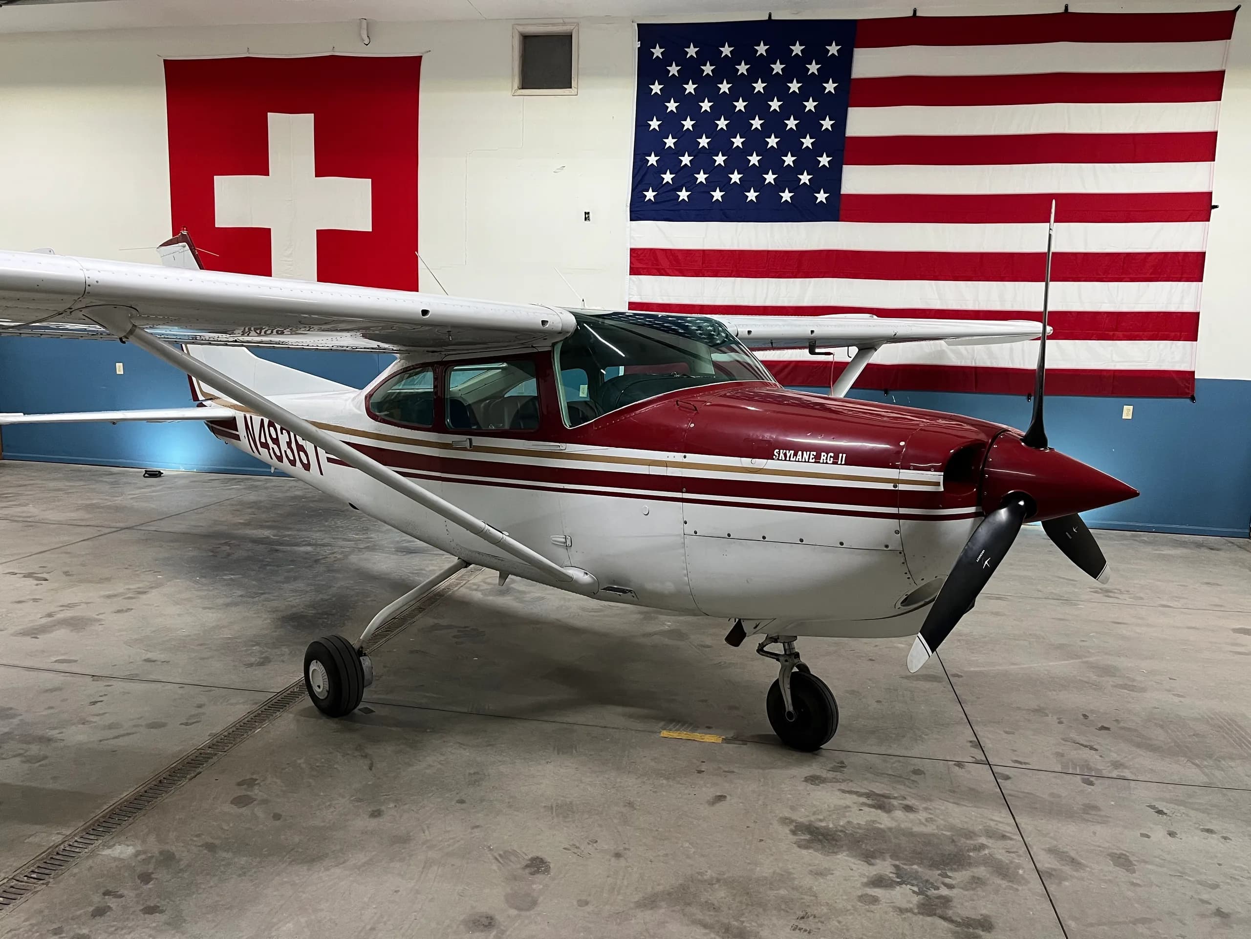 A red and white Cessna-like aircraft, parked in a hangar with Swiss and American flags.