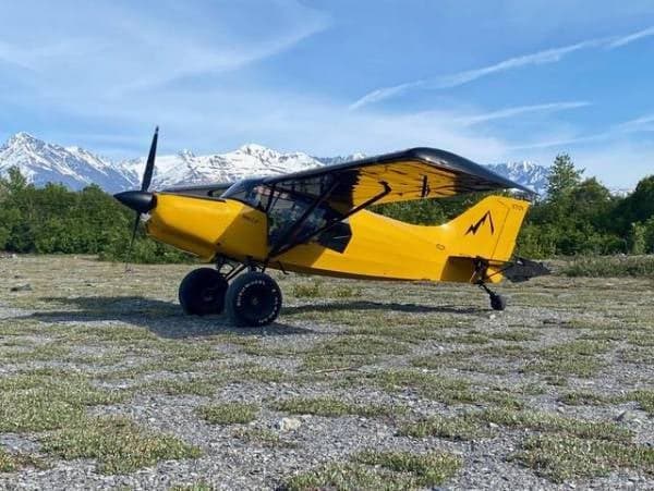 A yellow Maule aircraft is parked in a field with mountains in the background.