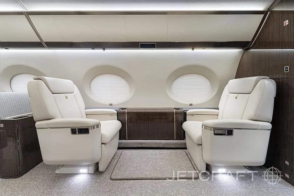 Two cream leather executive seats with foot lighting in a private jet cabin, facing a dark wood cabinet.