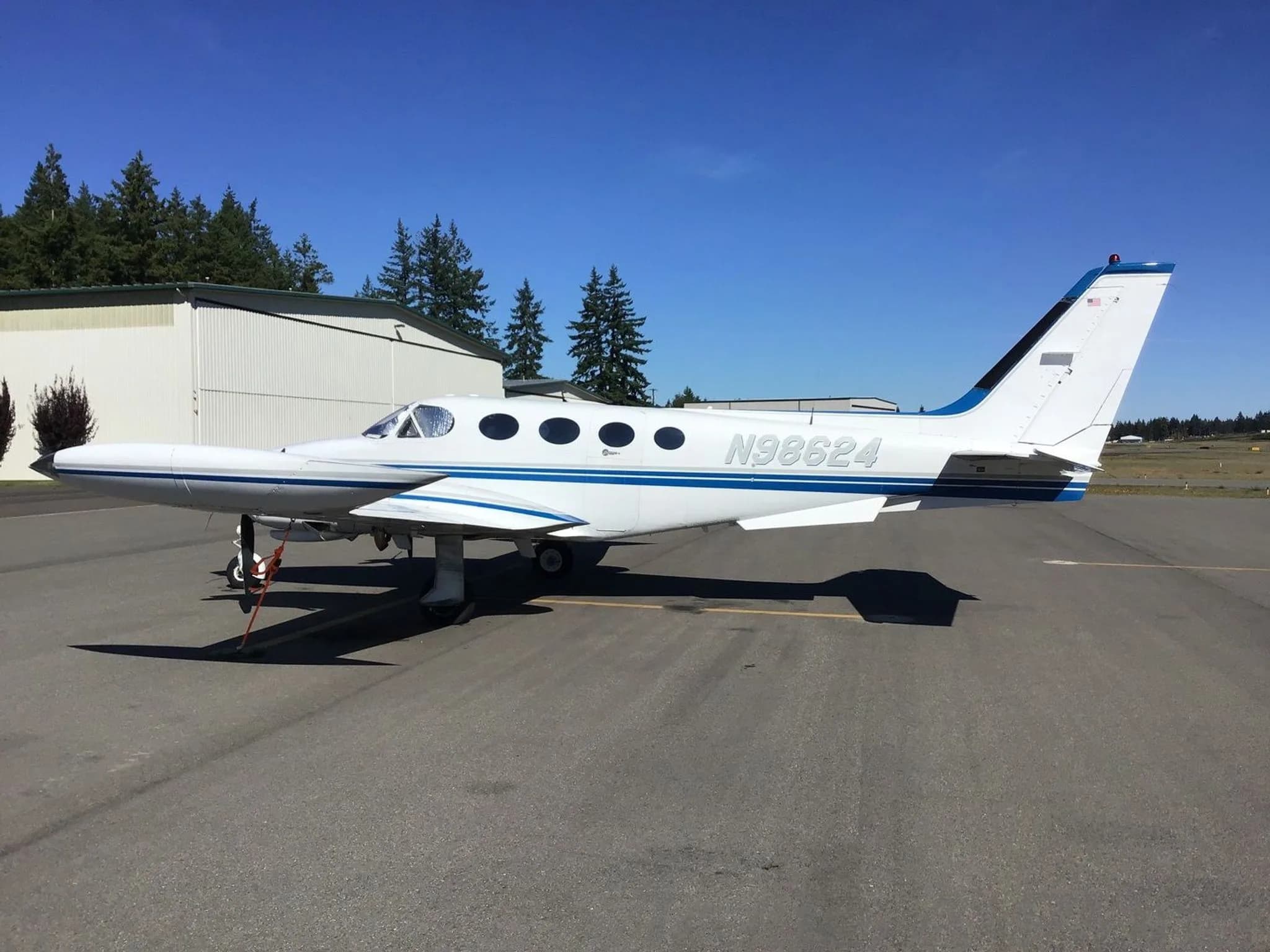 A white and blue twin-engine Cessna 340 aircraft, N98624, parked on an airfield with hangars and trees in the background.