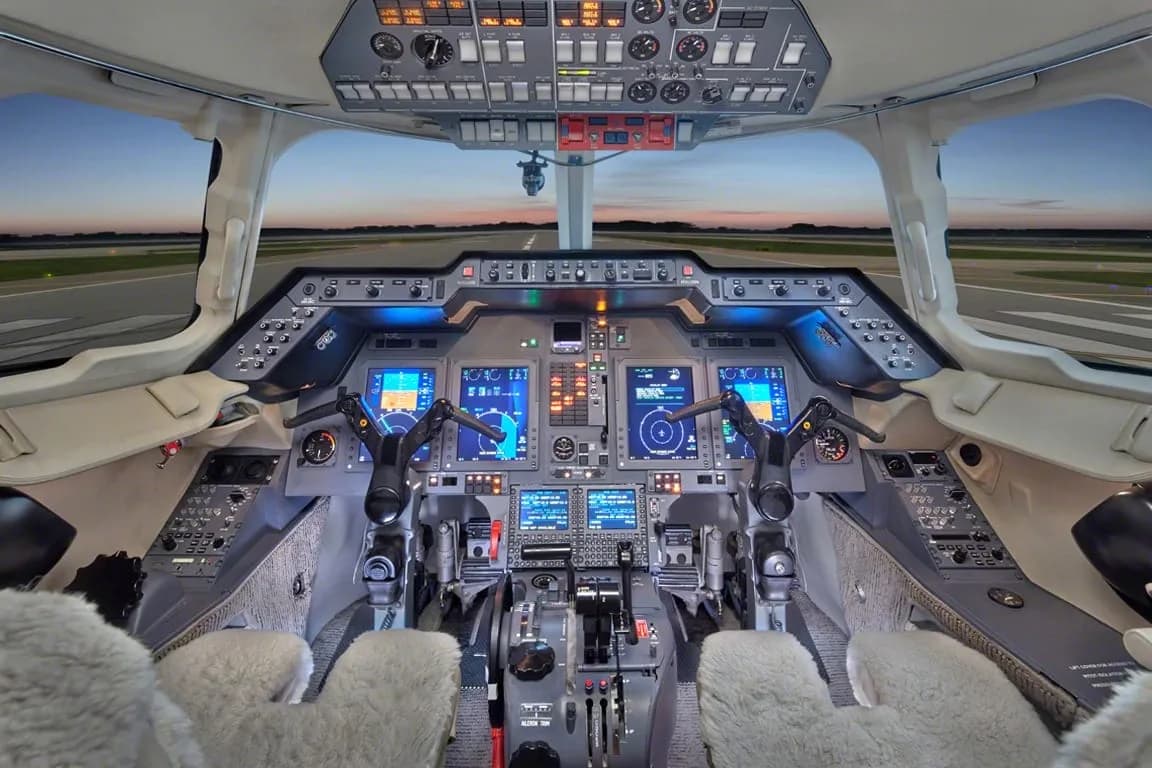 The modern cockpit of a private jet with multiple digital displays and controls, overlooking a runway at dusk.