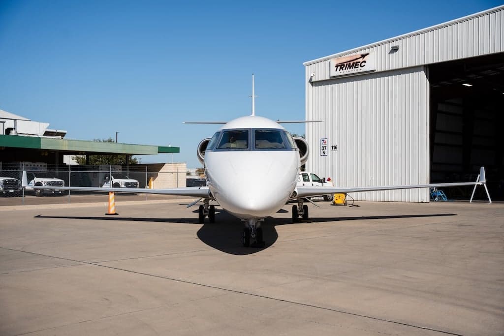 A white business jet, seen head-on, parked on the tarmac in front of a Trimec Aviation hangar.