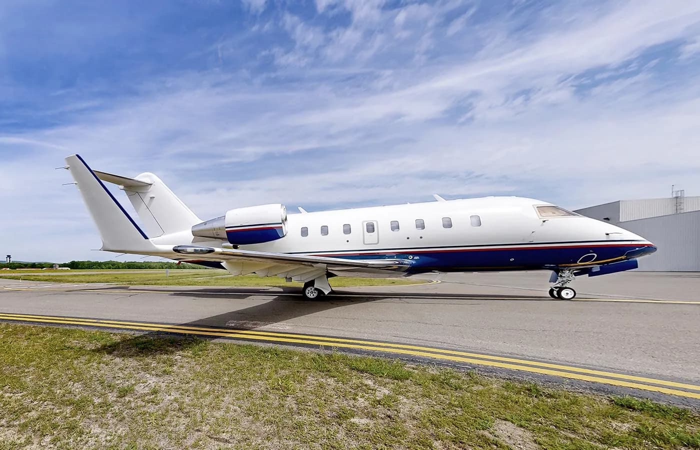 A white and blue Bombardier Challenger business jet is parked on a tarmac under a partly cloudy sky.