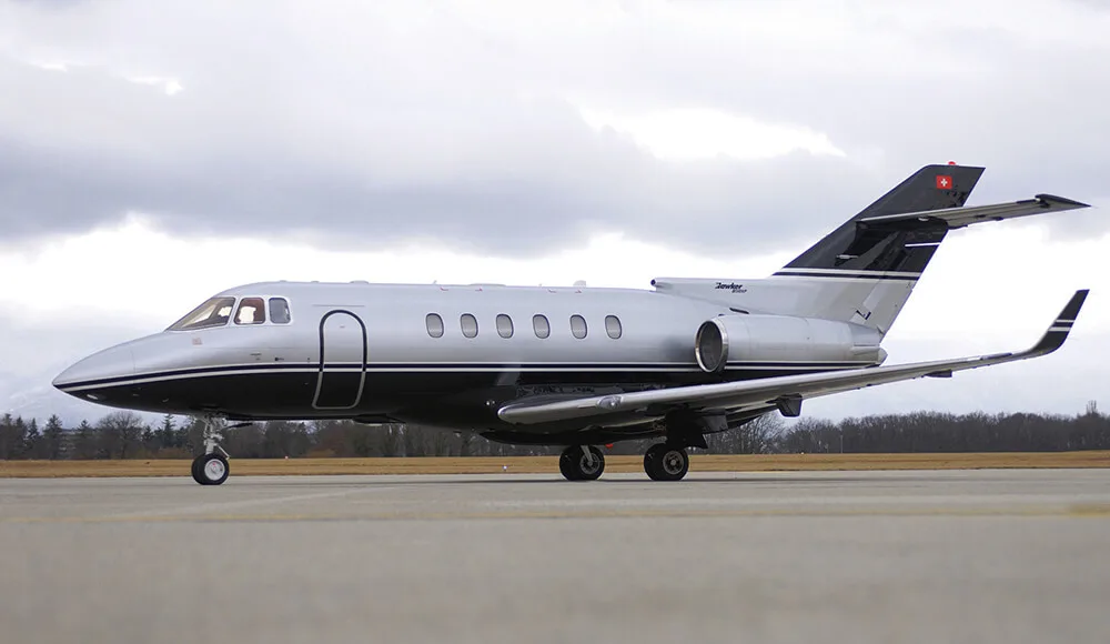 A silver and black Hawker 800XP business jet, with a Swiss flag on its tail, parked on an airfield under a cloudy sky.