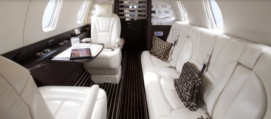 The spacious interior of a private jet features a mix of white leather seating, including a sofa and individual chairs, with dark wood accents.