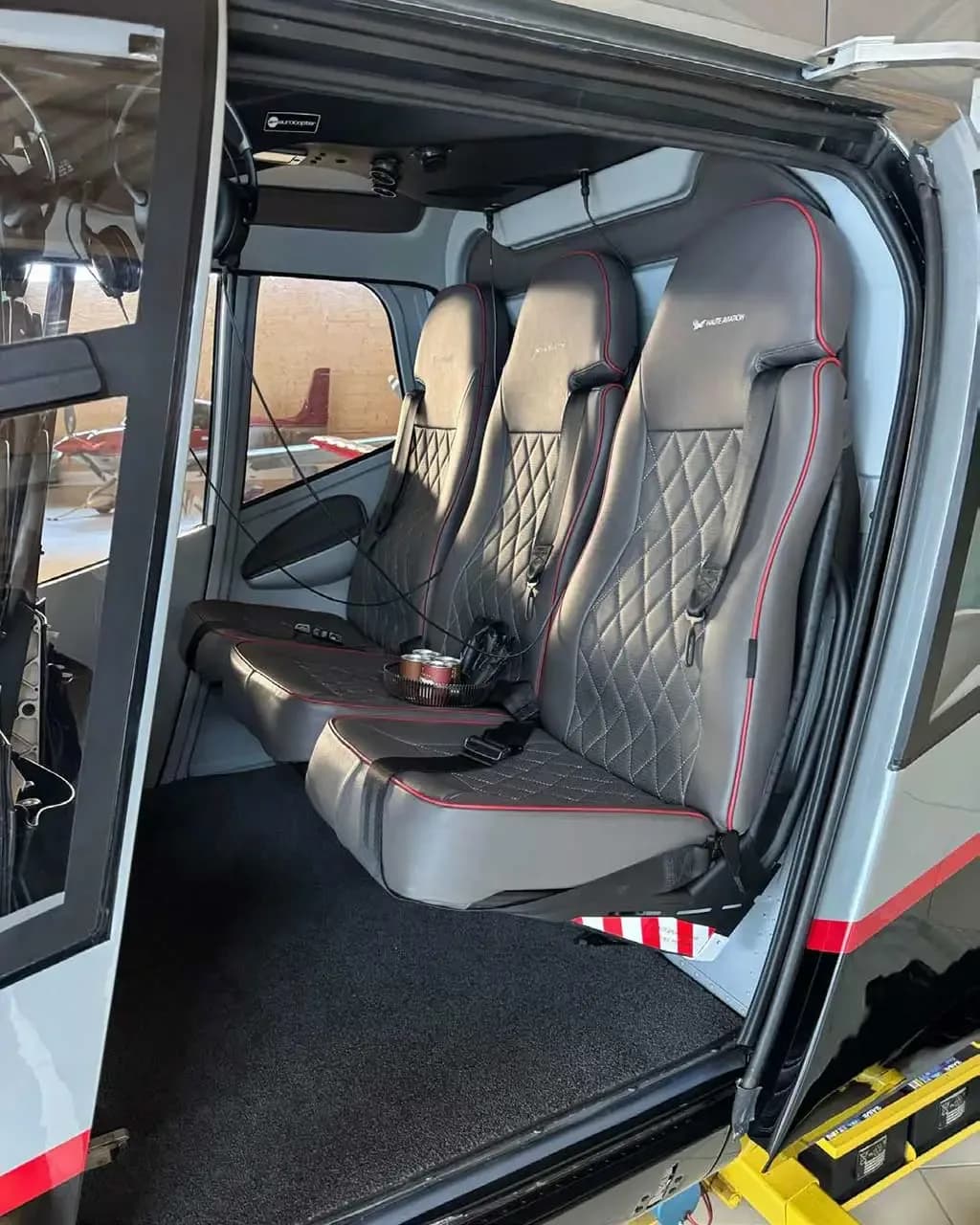 The interior of a helicopter showing three grey quilted passenger seats with red stitching and an open door.