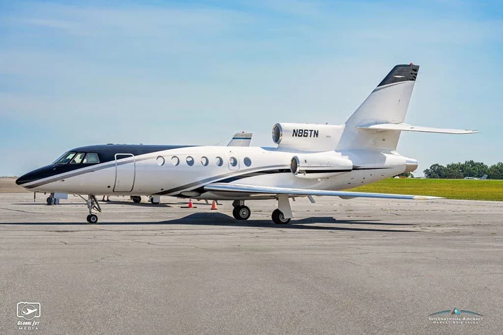A Dassault Falcon 900EX business jet, registration N86TN, parked on an airfield, showing its black nose and white fuselage with a grey stripe.
