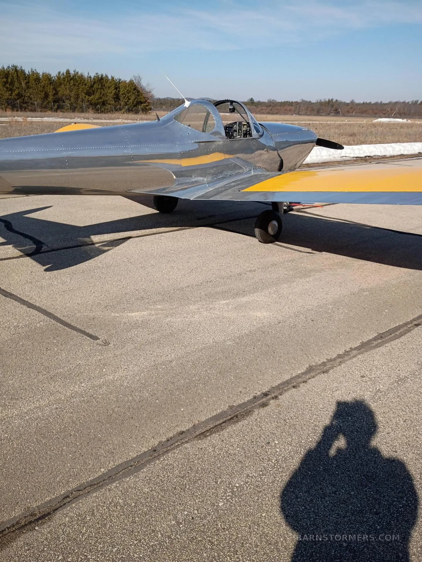 A side view of a silver aircraft with yellow wings on an airfield, with a pilot's shadow in the foreground.