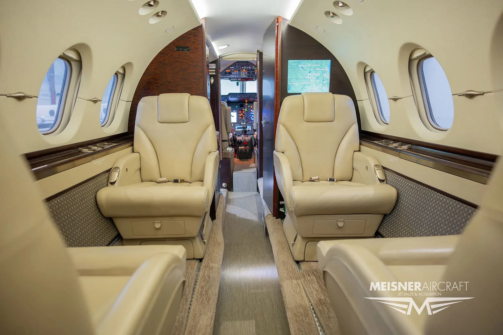 The luxurious cabin interior of a business jet, featuring beige leather seats and a view towards the cockpit.