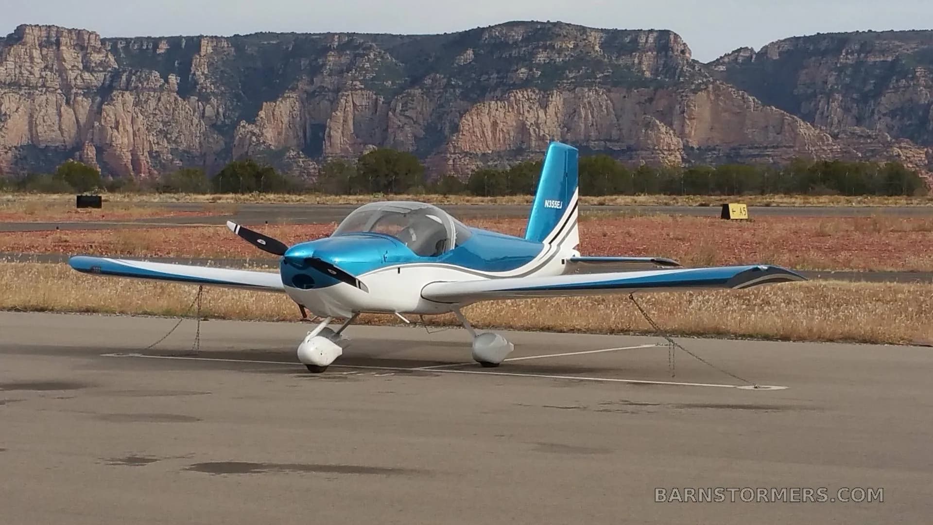 A blue and white single-engine aircraft with registration N355EJ is parked on an airfield, viewed from the front with mountains behind it.