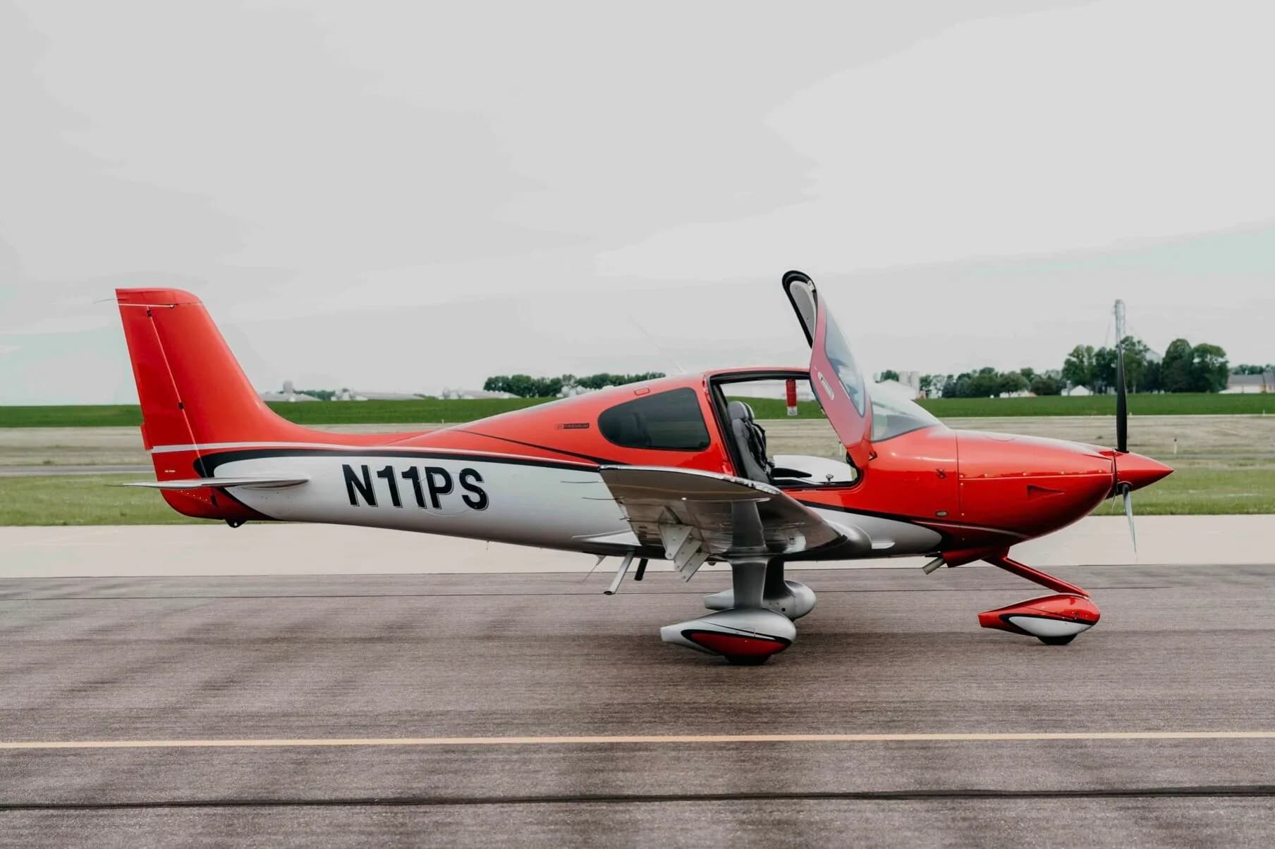 A red and silver Cirrus SR20 Premium aircraft (N11PS) with the pilot's door open, parked on an airfield tarmac.