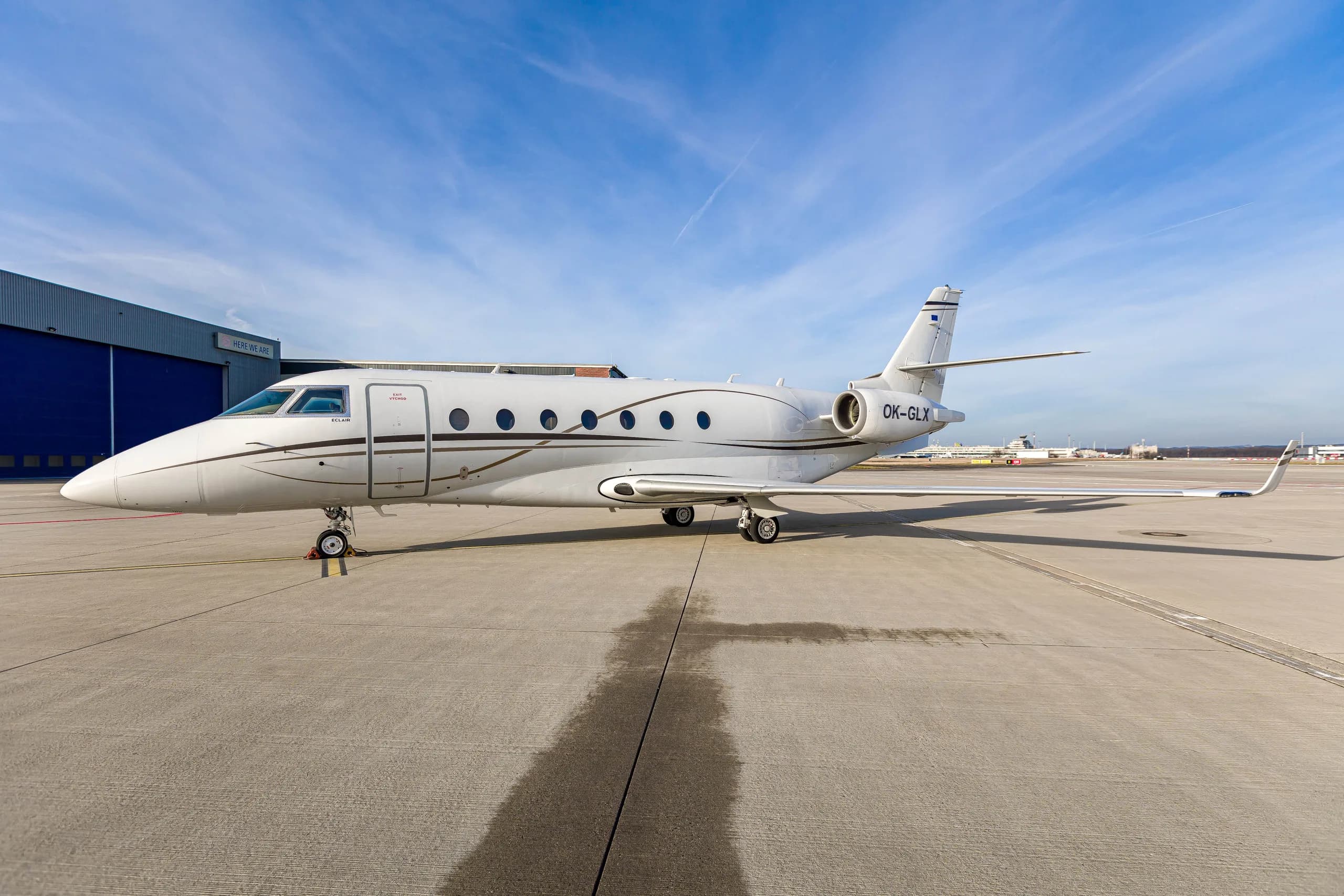 A white Gulfstream G200 business jet with registration OK-GLX parked on an airport tarmac under a blue sky.