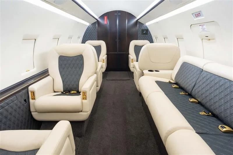 The interior of a private jet cabin featuring cream and dark grey quilted seats and a lounge sofa.