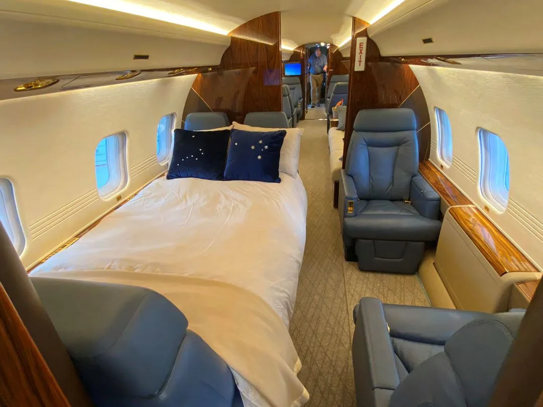 The cabin interior of a business jet shows a bed made up with white linens and blue pillows, alongside blue leather seats.