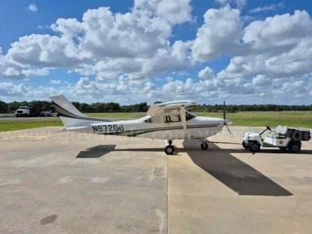 A white Cessna 206 aircraft with green stripes, registration N972GJ, parked on an airfield ramp under a partly cloudy sky.