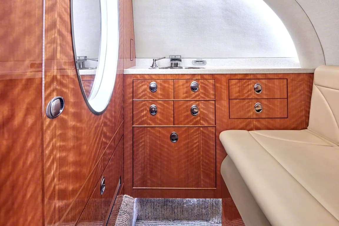 A close-up view of the aircraft interior showing polished wood cabinetry, a small sink, and a beige seat.