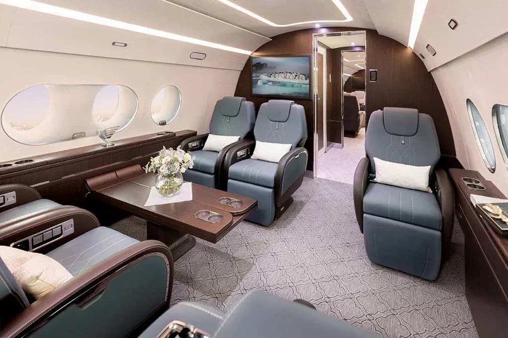 A private jet cabin featuring blue leather seats, a wooden table with flowers, and a TV screen displaying penguins.