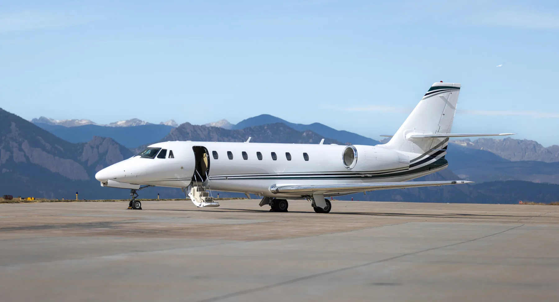 A white business jet with green stripes parked on an airfield with its door open, set against a backdrop of mountains.