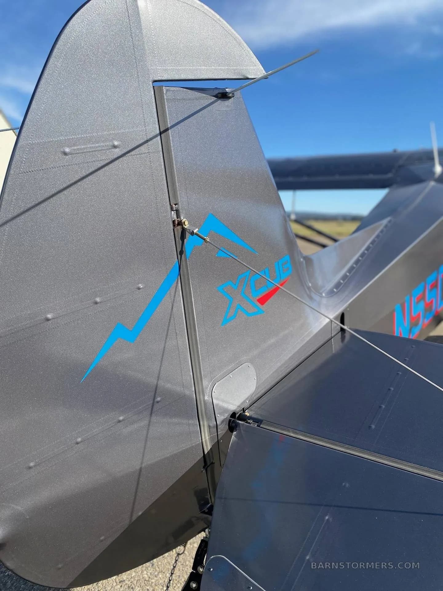 A close-up view of the tail of a dark gray X-Cub aircraft, showing the blue 'XCUB' logo and registration N55CX.