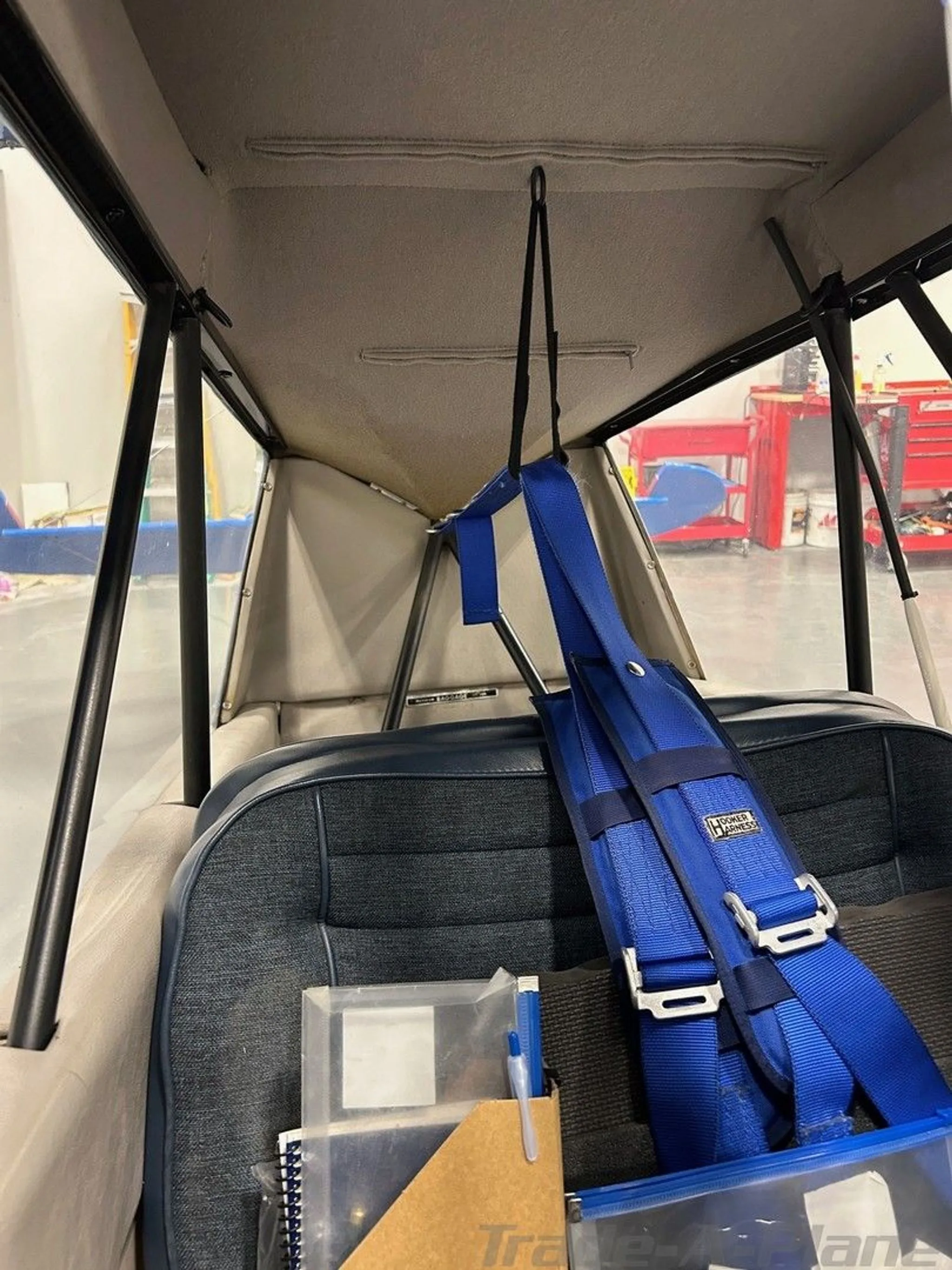 The rear interior of an aircraft cabin, showing a blue seat with a harness and the overhead structure.