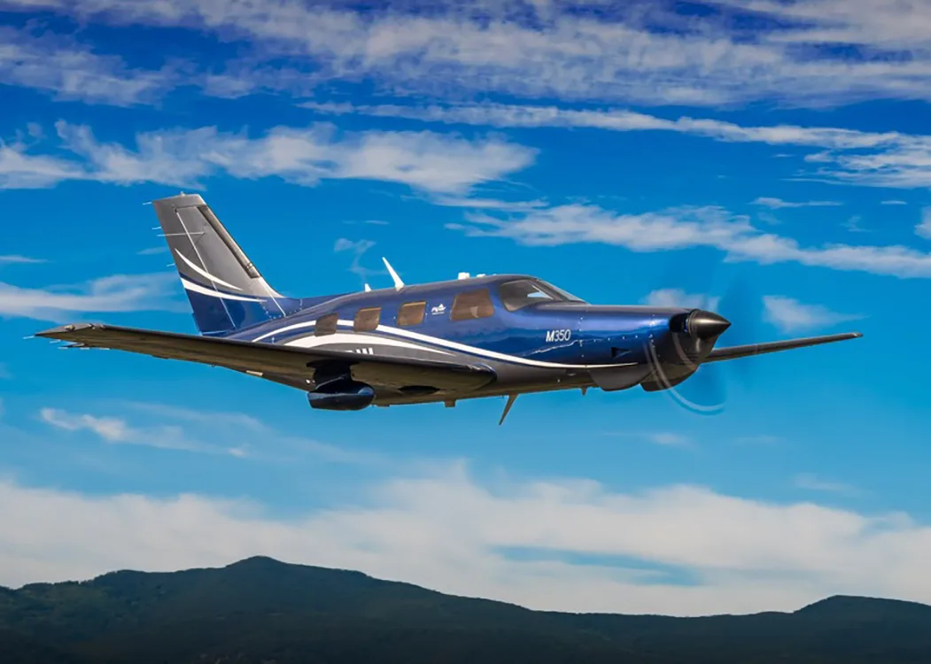 A dark blue Piper M350 aircraft with white and grey accents flying over a mountainous landscape.