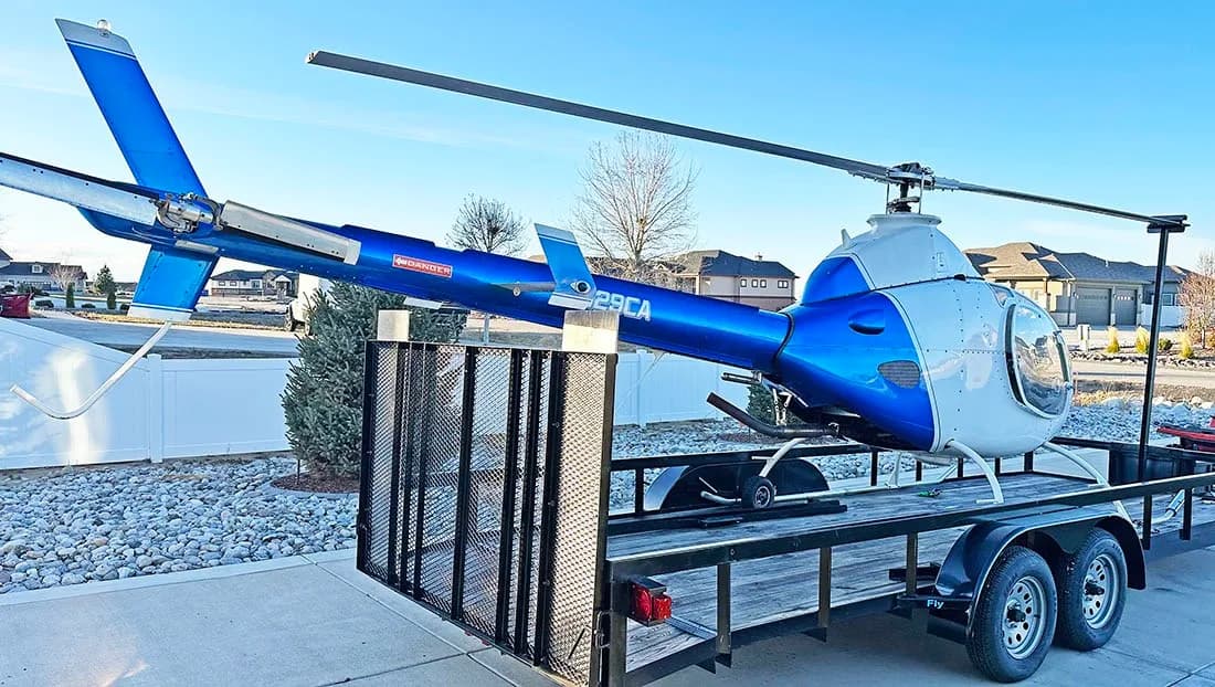 A blue and white helicopter (N129CA) on a black trailer, viewed from the rear-left, parked in a residential area.