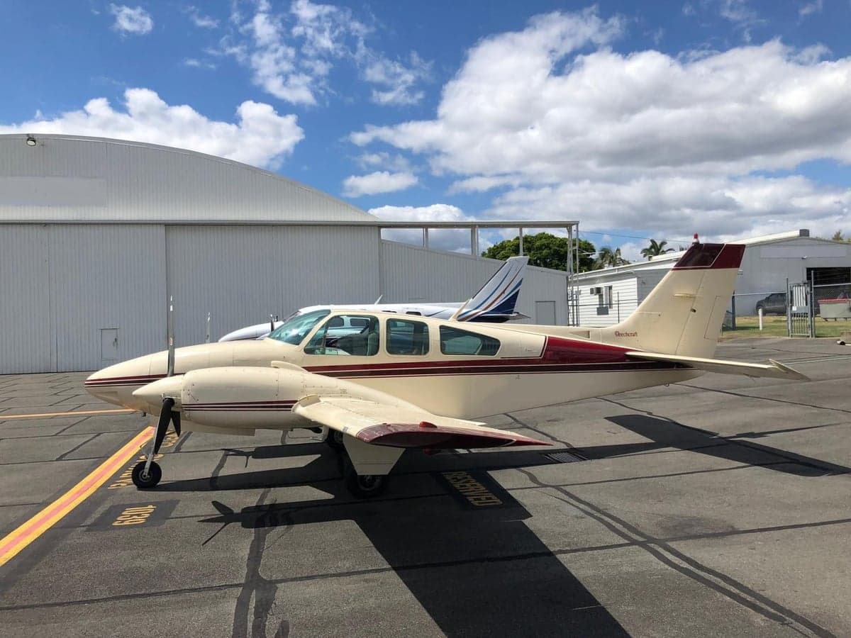A cream and maroon Beechcraft Baron aircraft parked on an airport tarmac next to a white hangar.
