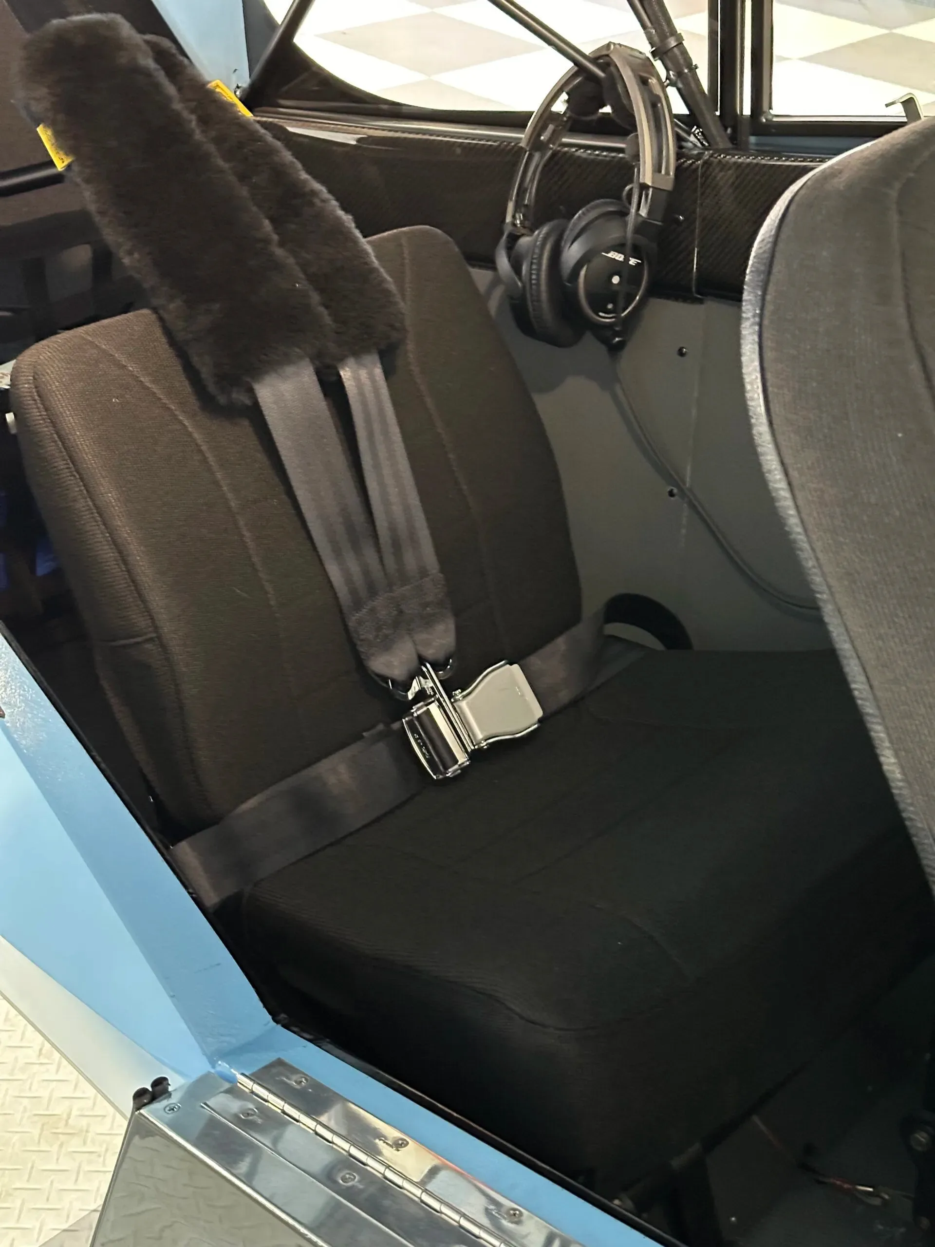 A close-up of a black fabric aircraft seat with a sheepskin-covered shoulder harness and a seatbelt buckle.