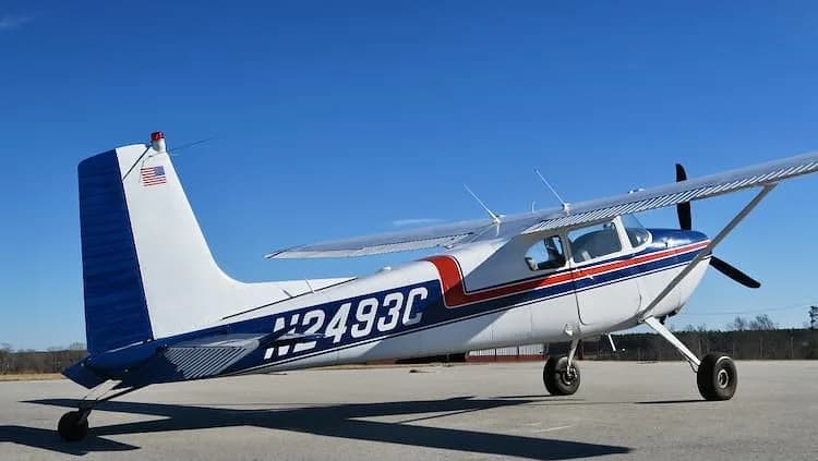 A side view of a white and blue Cessna 180 Skywagon with red stripes, registration N2493C, parked on an airfield.