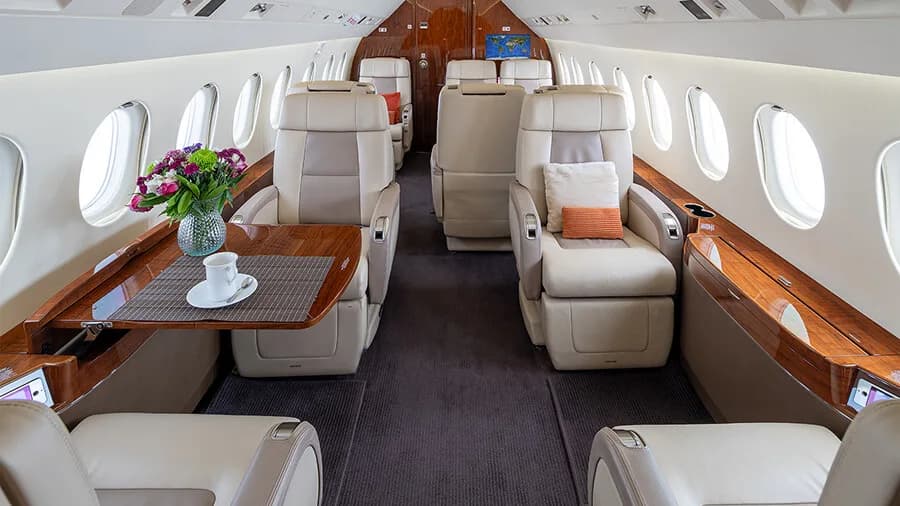 A wide view of the private jet cabin with multiple beige leather seats, polished wood tables, and large windows.