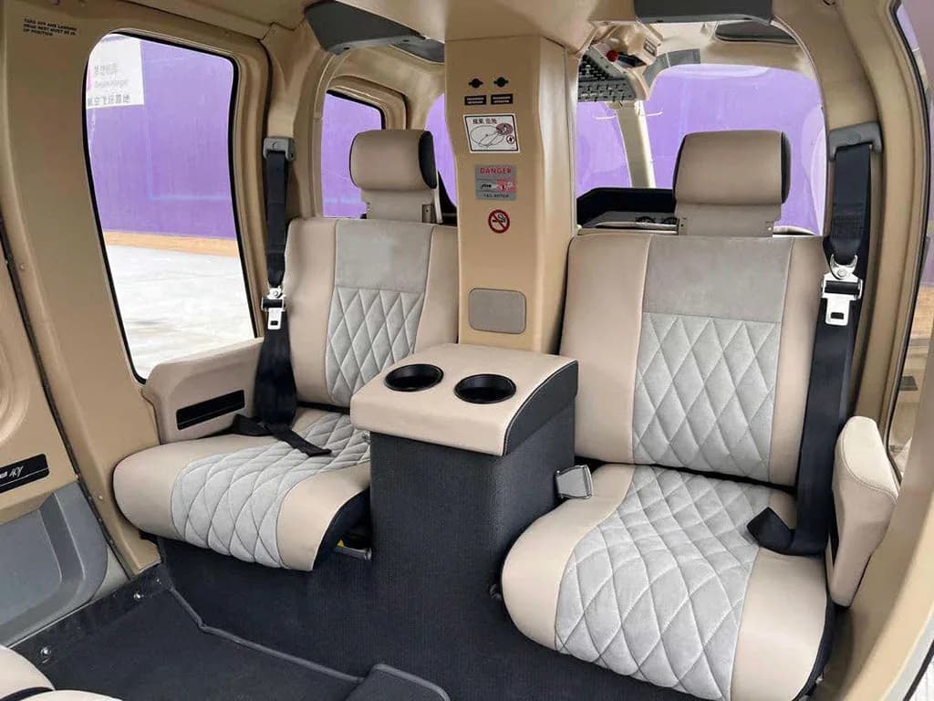 The rear cabin interior of a helicopter, showing two beige and gray quilted seats with a central armrest and cup holders, viewed from the front.