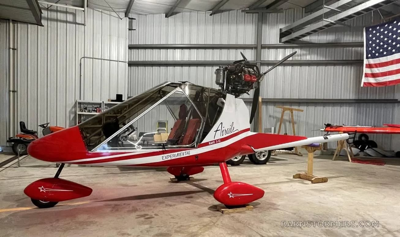 A red and white RANS S-12 Airaile experimental aircraft with a pusher propeller, parked in a hangar.