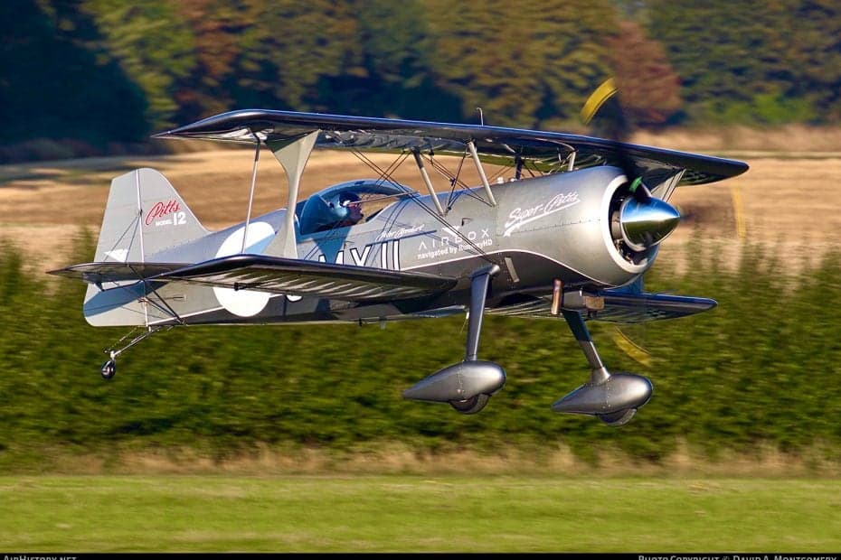 A silver biplane in flight over a green field.