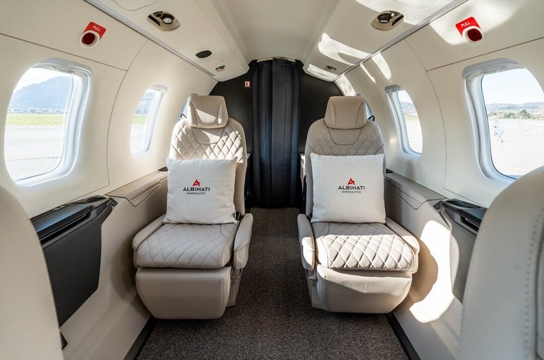 A view of the private jet cabin interior showing two forward-facing quilted leather seats with 'ALBINATI AERONAUTICS' branded pillows.