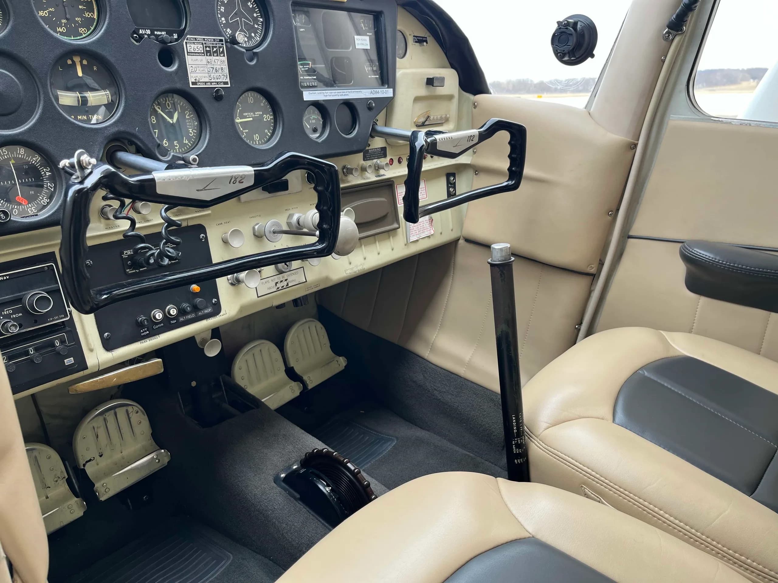 The cockpit of a Cessna 182 aircraft, showing the instrument panel, dual yokes, and rudder pedals.