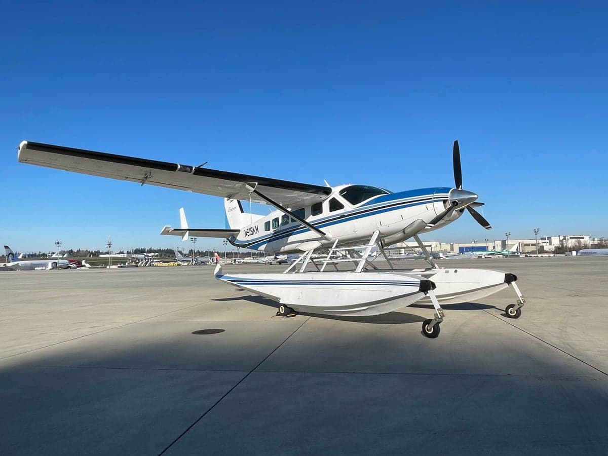 A white and blue Cessna Caravan on floats, parked on an airfield under a clear blue sky.