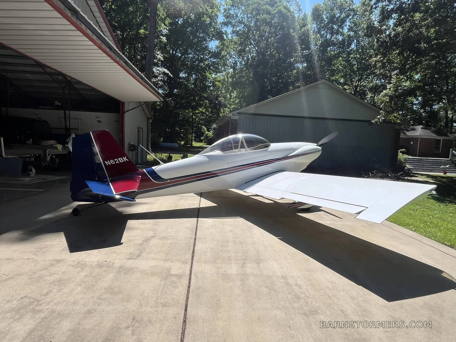 A white single-engine aircraft with red and blue tail stripes, parked on concrete in front of an open hangar, with sunlight shining through trees.
