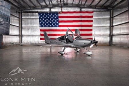 A silver single-engine aircraft with its doors open is parked in a hangar with a large American flag in the background.