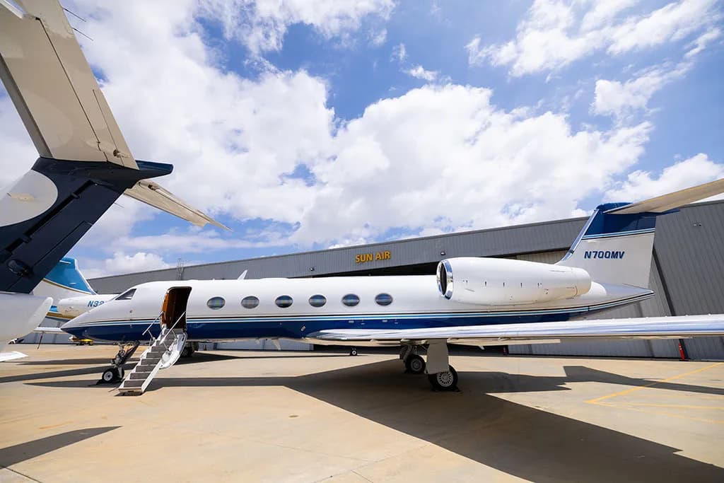 A white Gulfstream G550 with blue stripes, tail number N700MV, parked on an tarmac with stairs deployed, in front of a hangar.