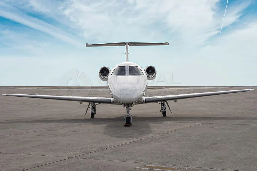 A head-on view of a white Embraer Phenom 300 business jet parked on an airfield under a blue sky.