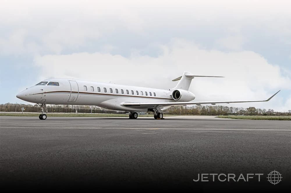 A white Bombardier Global business jet with brown accent stripes parked on an airfield under a cloudy sky.