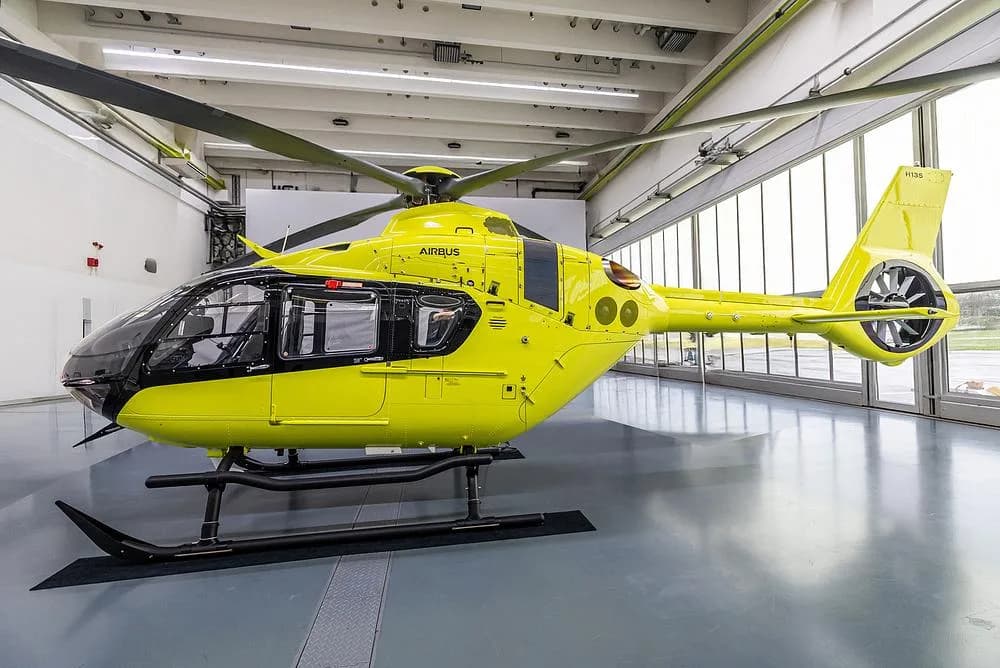 A bright yellow Airbus H135 helicopter with black accents is parked in a hangar, viewed from the front-right side.