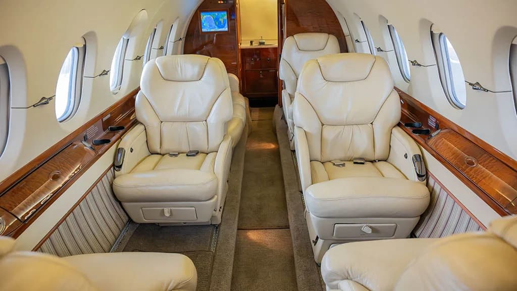 The interior cabin of a private jet with two rows of cream leather seats facing each other, and wood paneling.